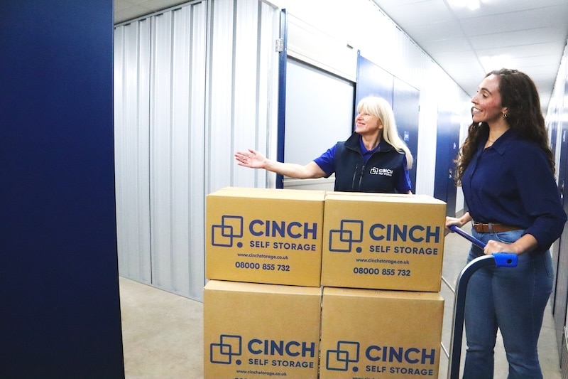 Storage Exeter. Image shows a Cinch Storage Employee showing a female customer a storage unit. The female customer is pushing a trolley loaded with Cinch Self Storage cardboard boxes.
