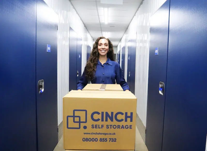 Storage in Exeter. Image shows a woman pushing a trolley filled with Cinch Self Storage cardboard boxes down a storage unit corridor.