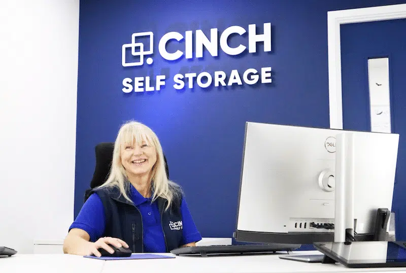 Storage rooms Exeter. Image shows a Cinch Self Storage employee sitting at her desk smiling with the Cinch Self Storage logo behind her.